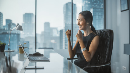Fototapeta premium Excited Businesswoman in Stylish Dress Sitting at a Desk in Modern Office, Using Laptop Computer. Successful Manager Ecstatic About Winning a Real Estate Investment Deal for Corporate Business.