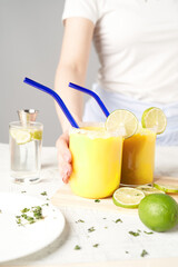 Cropped shot of a woman wearing light-colored clothes and standing by a table. The lady is holding a cocktail with a lime wedge and a blue glass straw. There is a plate, glasses and a lime next to it.