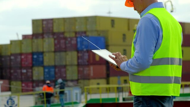 Sea port cargo ship technology. Middle aged taskmaster works on tablet while workers prepare metal containers to load into vessel in dockyard