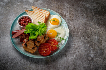 Serving a dish from the restaurant menu. Delicious and healthy breakfast with fried eggs, beans, sausages, grilled vegetables, tomatoes and mushrooms with salad on a plate against a gray stone table