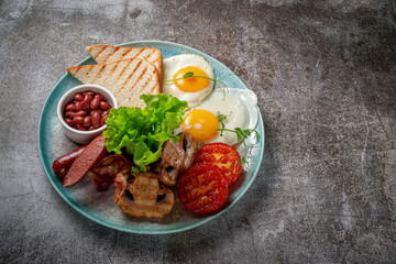 Serving a dish from the restaurant menu. Delicious and healthy breakfast with fried eggs, beans, sausages, grilled vegetables, tomatoes and mushrooms with salad on a plate against a gray stone table