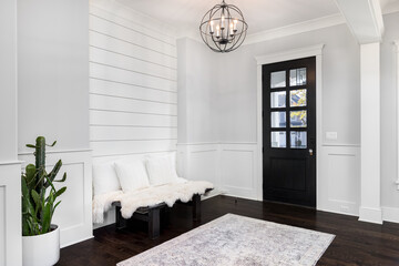 A beautiful foyer entrance with a light hanging above the dark hardwood floors, a bench in front of a shiplap wall, and a dark door with windows. - obrazy, fototapety, plakaty