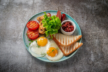 Serving a dish from the restaurant menu. Delicious and healthy breakfast with fried eggs, beans, sausages, grilled vegetables, tomatoes and mushrooms with salad on a plate against a gray stone table