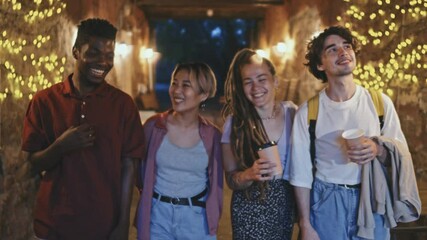 Tracking medium shot of group of young diverse friends walking towards camera along narrow road decorated with yellow lights at night and having conversation