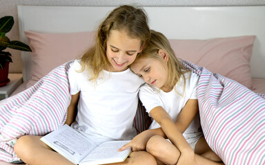 two cute little girls sisters are sitting on the bed covered with a warm blanket and reading a...
