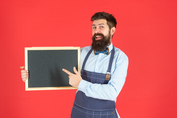 Happy guy in barber apron smile pointing finger at blackboard red background copy space, barbershop