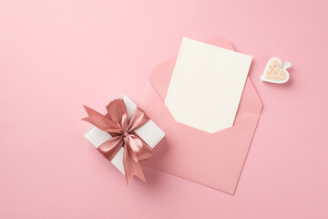 Top view photo of white giftbox with pink silk ribbon bow decorative heart and open pink envelope with card on isolated pastel pink background with copyspace