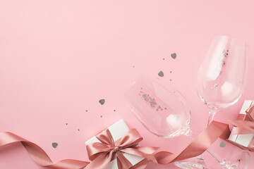 Top view photo of white gift boxes with pink silk ribbon bows two wineglasses silver sequins and heart shaped confetti on isolated pastel pink background with copyspace
