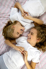 Obraz na płótnie Canvas two blonde girls sisters lie on the bed facing each other, laugh and rejoice, show with their hands a sign of heart, love and harmony within the family, view from above
