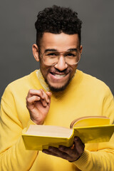 sly african american man in glasses and yellow sweater holding book and smiling isolated on grey.