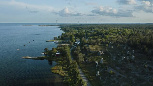 Aerial shot over a small costal community next to rauk stones with wind power plants in the distance