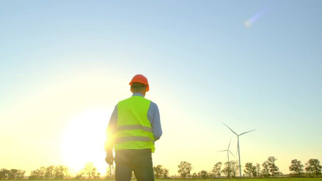 Wind turbines generate electricity in field. Builder in uniform walks at construction site of rural substation holding tablet backside view