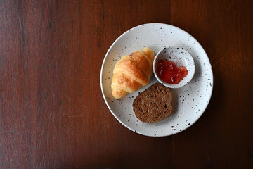 Top view of small croissant homemade cocoa bread with sweet strawberry jam on a plate on a wooden table. It is often eaten with hot beverages such as coffee, cocoa or fresh milk.
