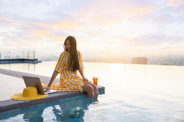Confident woman relaxing sitting in yellow summer dress sitting on the edge dangling her feet in swimming pool on skyscraper. Woman using social media on laptop for relax during sunset. Copy space
