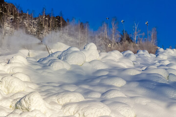 Snowdrifts and flying snow from a snow cannon on the mountainside against the background of trees and blue sky in winter