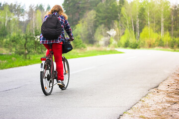 Naklejka premium Kiev, Ukraine. May 1, 2020. A girl, young woman in checkered shirt, red pants, black backpack riding bicycle on a country paved road in woods, park in spring, summer. Journey alone, healthy lifestyle.