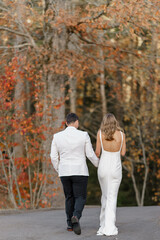 Fototapeta premium Newlyweds walking through the forest with their backs to the camera Bride in white dress and groom in a white suit walk along the downhill road between the trees Forest background Back view 