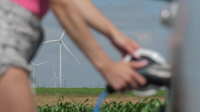Wind turbines generate power in corn field. Young woman takes plug off white e-car and closes cap at charging station on cloudy summer day closeup