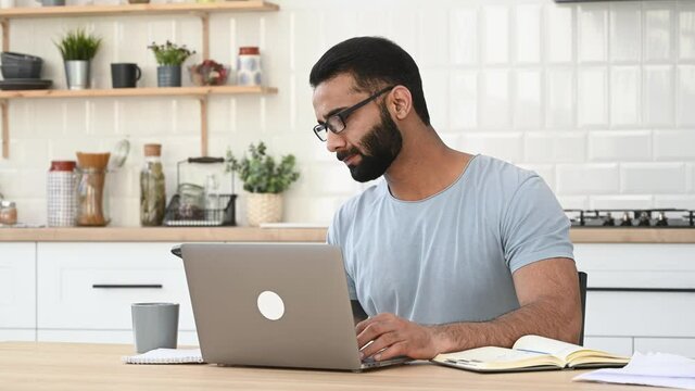 Busy successful Indian man, IT specialist or freelancer, sitting at work desk at home, having a nice cell phone conversation during work time, smile friendly, looks away. Mobile phone communication