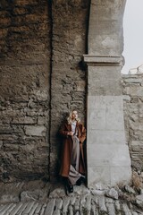 Stylish blonde girl in brown trousers, coat, suspenders, white shirt posing in an old stone tunnel. Fashionable young woman with hat.