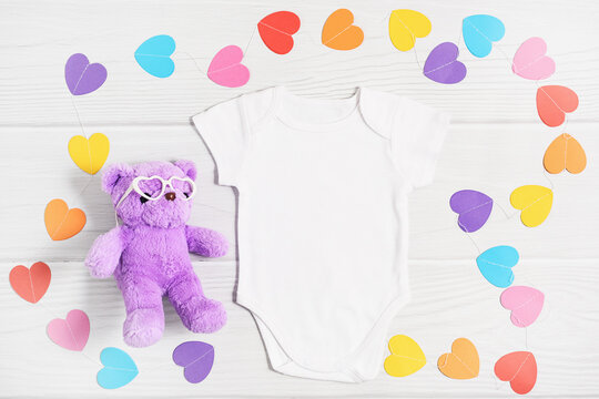 Mockup white baby shirt with paper colorful hearts. Happy Valentines Day baby apparel flatlay on white wood background, flat lay, top view, copy space.