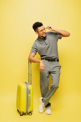 full length of stylish african american man in sunglasses and grey tennis shirt standing near luggage on yellow.