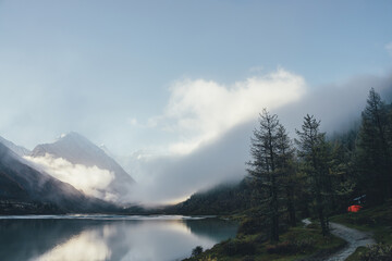 Atmospheric alpine landscape with coniferous trees near mountain lake and high snow-covered mountain in low clouds. Beautiful low lighting scenery with peaked top in thick fog and larches near lake.