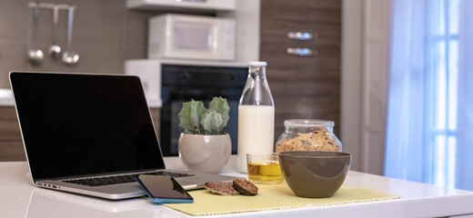 Table set for breakfast, kitchen in the background. Natural meal and technology devices. Interior view of a cozy and modern home. Millennial, youth, home working, home schooling, remote work concept.