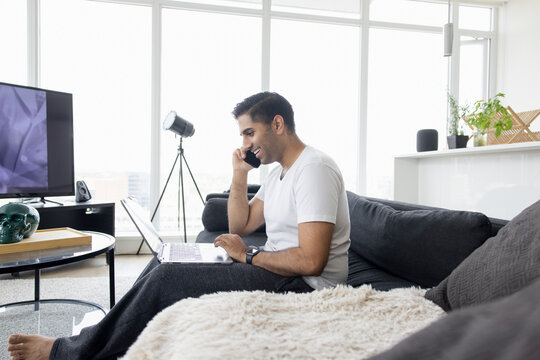 Man talking on smart phone and working at laptop on sofa