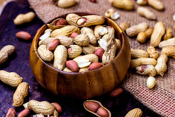 Raw unpeeled peanut nuts in nutshell in brown round wooden bowl on black metal and textile background.