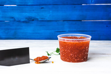 red caviar in a plastic jar on a white wooden background, next to a spoon with superimposed caviar,...