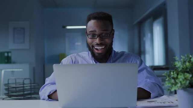 Young African American man in glasses working at laptop late celebrating success