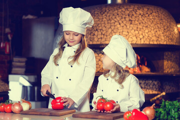 Little chefs in kitchen uniforms cut fresh vegetables with a knife.