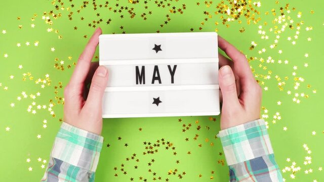 The hands of a young girl in a plaid shirt are holding a white board with the inscription May on a green background with shiny stars