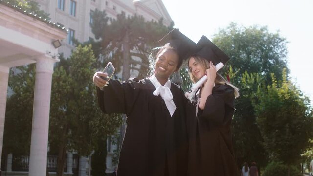 In the college garden amazing two best friends ladies students graduates posing with the smartphone taking pictures they wearing suits and caps. Shot on ARRI Alexa Mini.