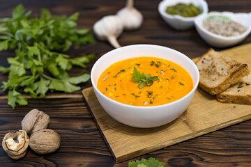 pumpkin cream soup in a white bowl with herbs, seeds, garlic and walnuts on a dark wooden...