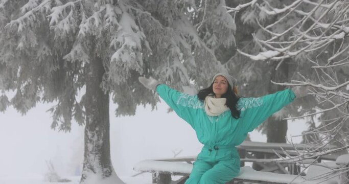 Young in winter clothes woman in full height outdoors. Adorable young adult girl in ski suit sitting alone on the bench and then raising hands in the mountain snowy forest.