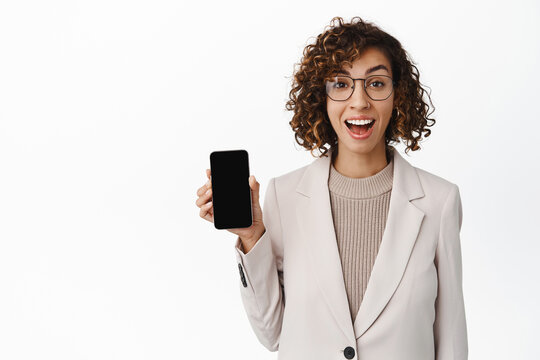 Enthusiastic corporate woman in glasses showing mobile phone application, empty smartphone screen, wearing beige suit, standing over white background