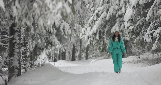 Young woman winter portrait in full height outdoors. Adorable young adult girl in ski suit walking in the mountain snowy forest.