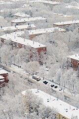 Snow-covered roofs of apartment buildings after snowfall the day before. Snow road between houses. Fluffy snow lies on branches of trees. Cars are parked along road