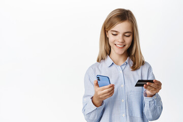 Excited blond teen girl, child showing credit card unior, looking happy at smartphone app screen, standing over white background, bank account for children concept