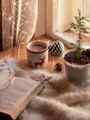 christmas still life. cup of cocoa, open book, fir cones, fir tree, white blanket, handmade snowflakes near the window
