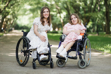 Two beautiful young women with disability sitting in wheelchairs and smiling on camera among green summer park. Female persons wearing stylish dresses with floral print.