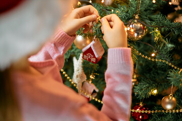 Close-up of a girl in a santa hat hangs a Christmas tree toy a house on a Christmas tree. Merry...