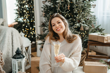 Happy young woman stretches out her hand with a glass of sparkling wine, looks at the camera, celebrates New Year's Christmas holidays, congratulates the family with a video call.