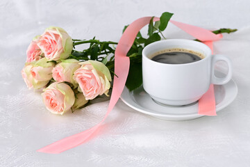 white cup of fragrant coffee, beautiful white roses and pink satin ribbon on white silk background. Valentine's day. Close-up