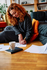 An artistic ginger girl is sitting on the floor in the morning and drawing portraits. She is enjoying her morning moments with a cup of fresh coffee.
