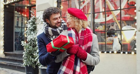 Obraz premium Man smiling and hugging his girlfriend and gives Christmas gift on decorated street. Christmas mood concept. Unexpected surprise.