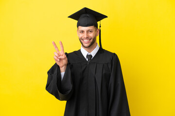 Young university graduate caucasian man isolated on yellow background smiling and showing victory sign