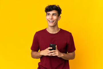 Young Argentinian man isolated on yellow background using mobile phone and looking up